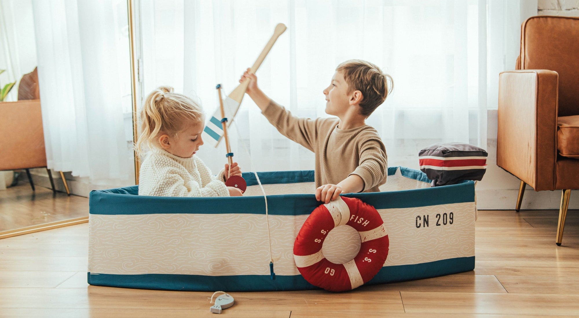 Two children playing with a toy boat in a room with a couch and window.