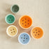 Colorful baby buttons in green, yellow, orange and blue arranged on neutral fabric for 9-12 month gift set
