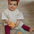 Smiling baby wearing red headband and burgundy leggings playing with orange sensory toy from baby gift set