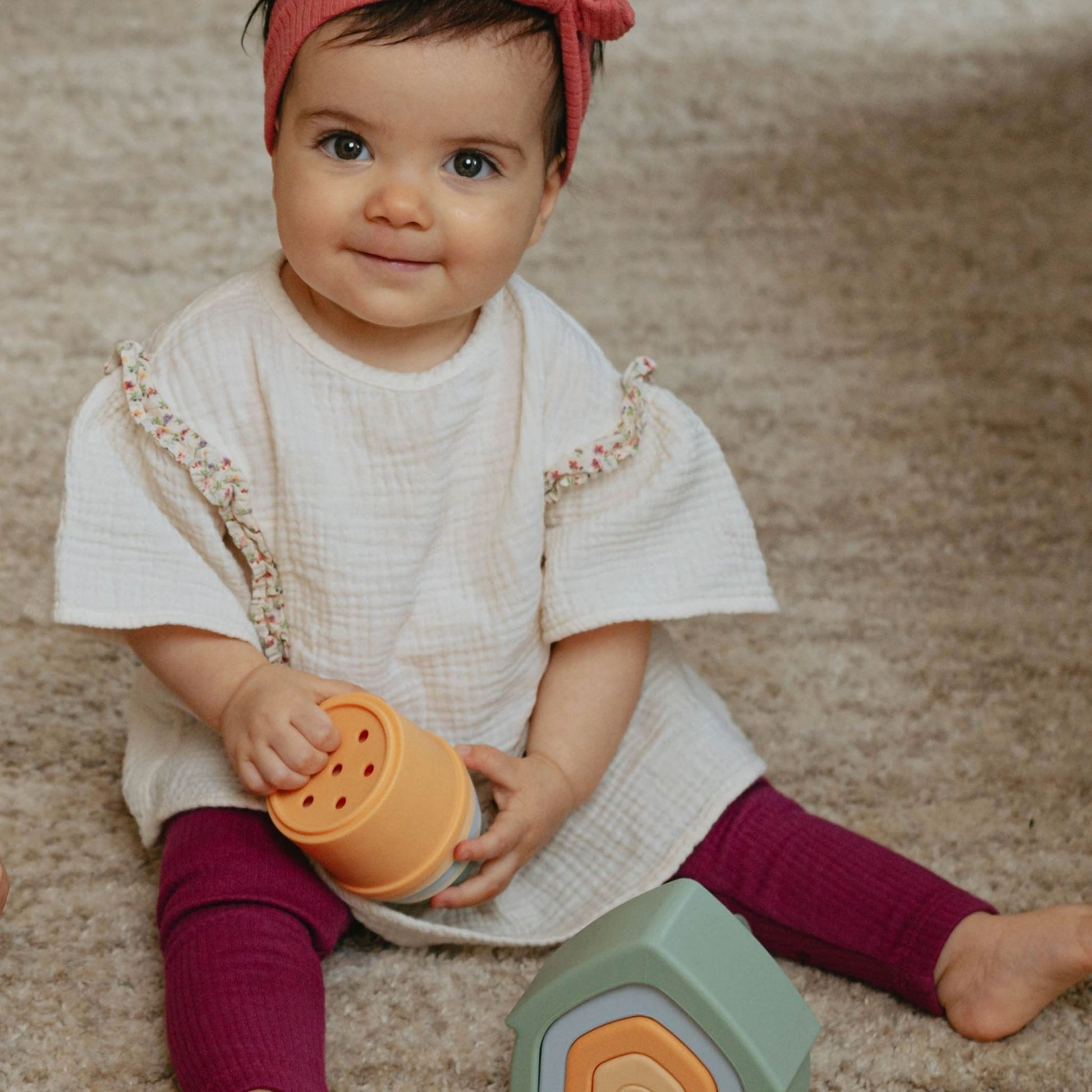 Smiling baby wearing red headband and burgundy leggings playing with orange sensory toy from baby gift set