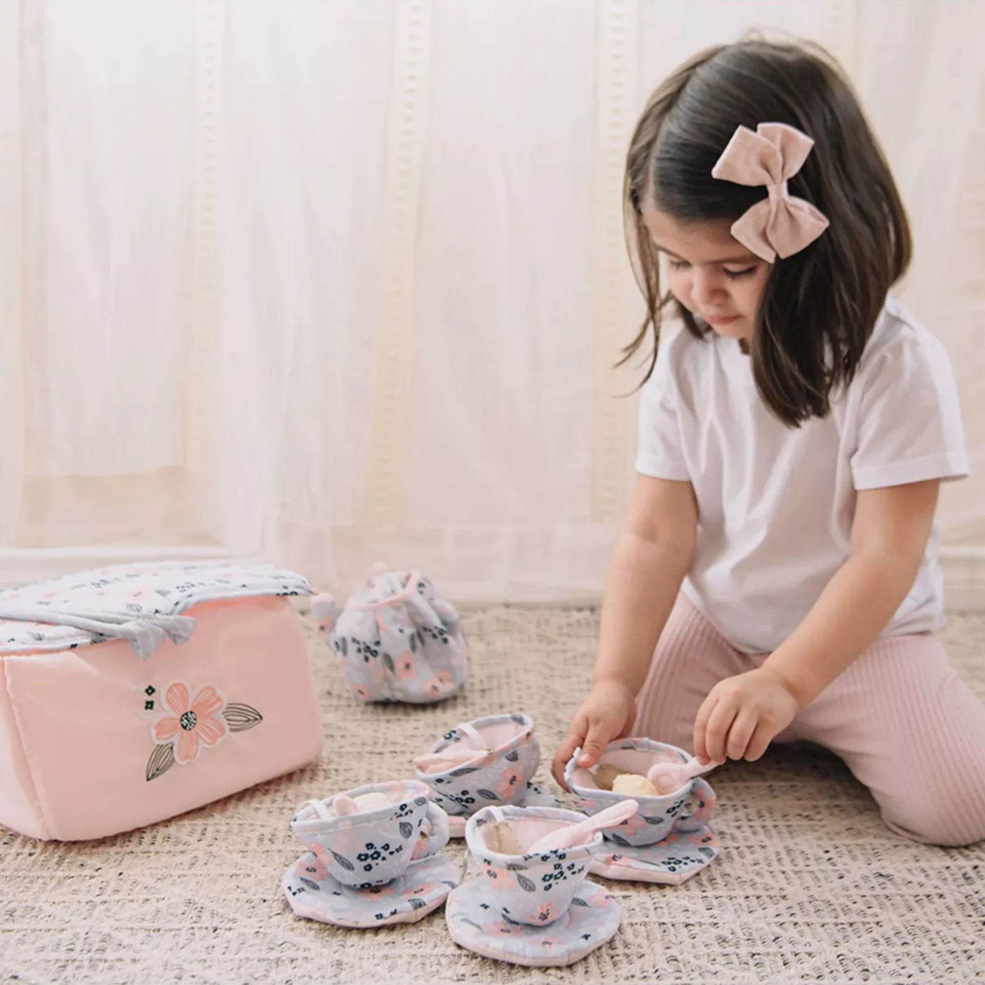 Young girl playing with plush tea set for toddlers in pink and floral pattern on floor