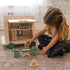 Child playing with a wooden toy house and furniture on a carpeted floor.