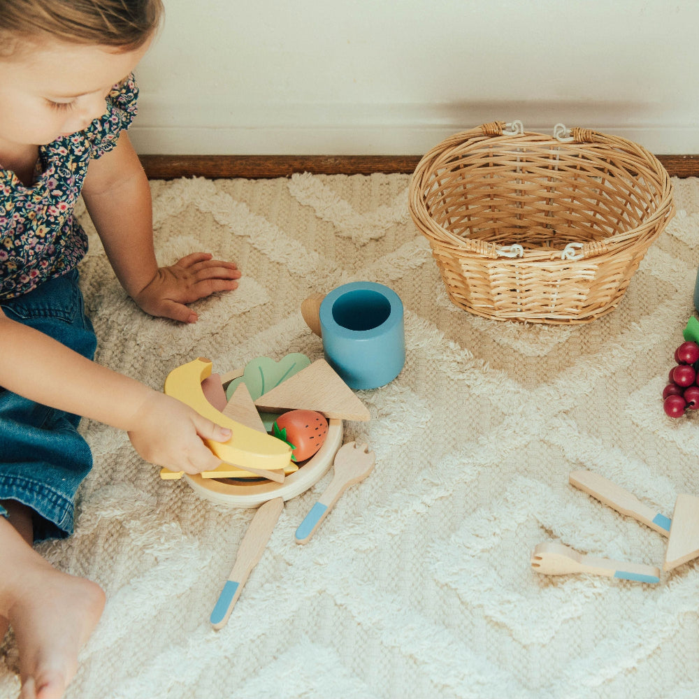 Child playing with wooden toy fruits and a basket on a carpeted floor.