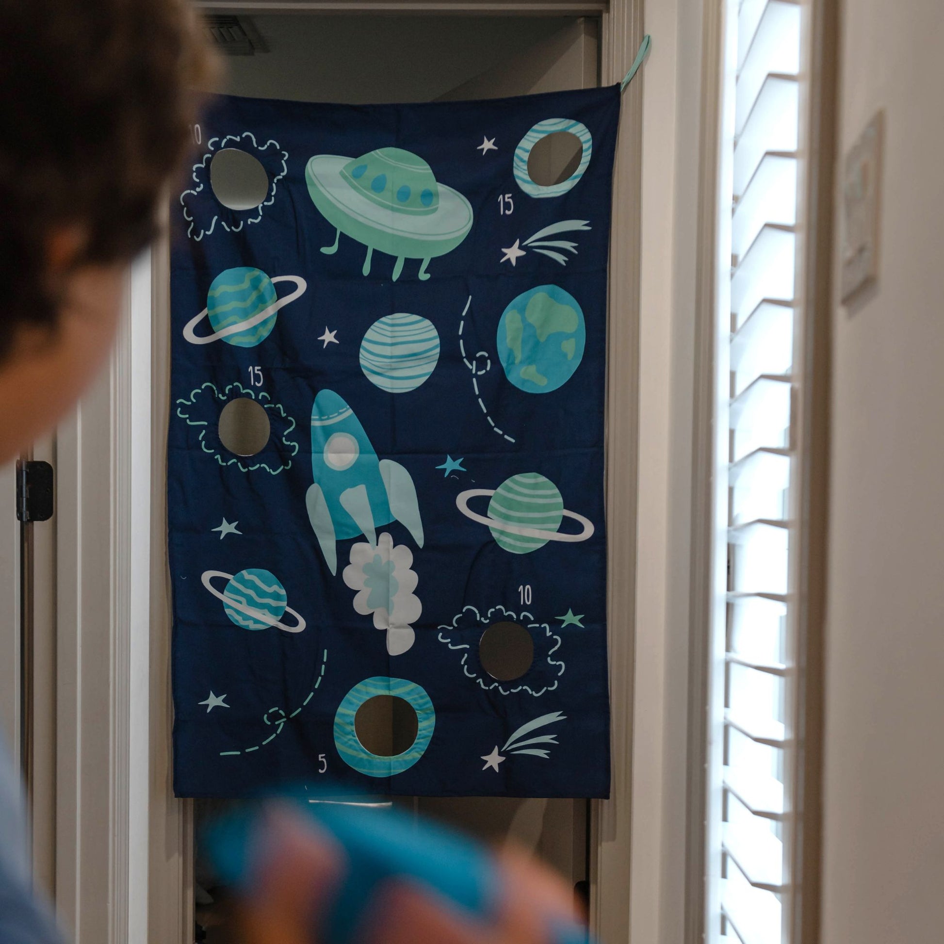 Space-themed doorway bean bag toss game with planets, rocket, and clouds on dark blue fabric