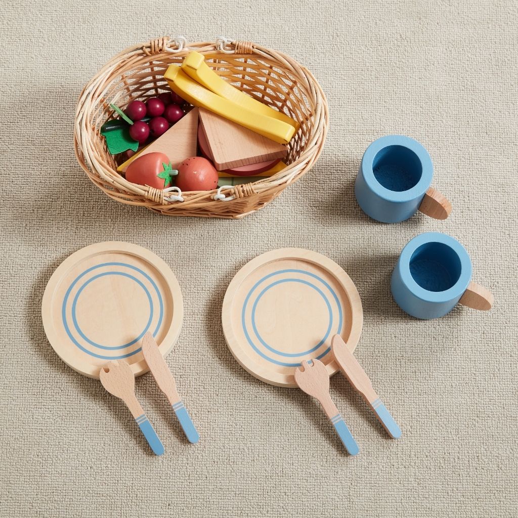 Set of wooden toys including plates, cups, and a basket with fruits on a beige surface