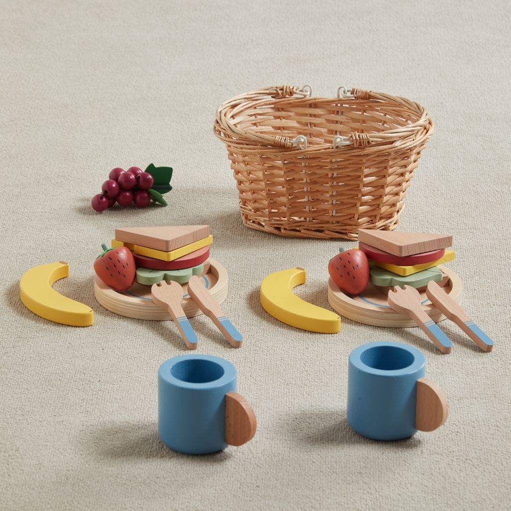 Wooden toy picnic set with basket, sandwiches, fruits, and cups on a neutral background