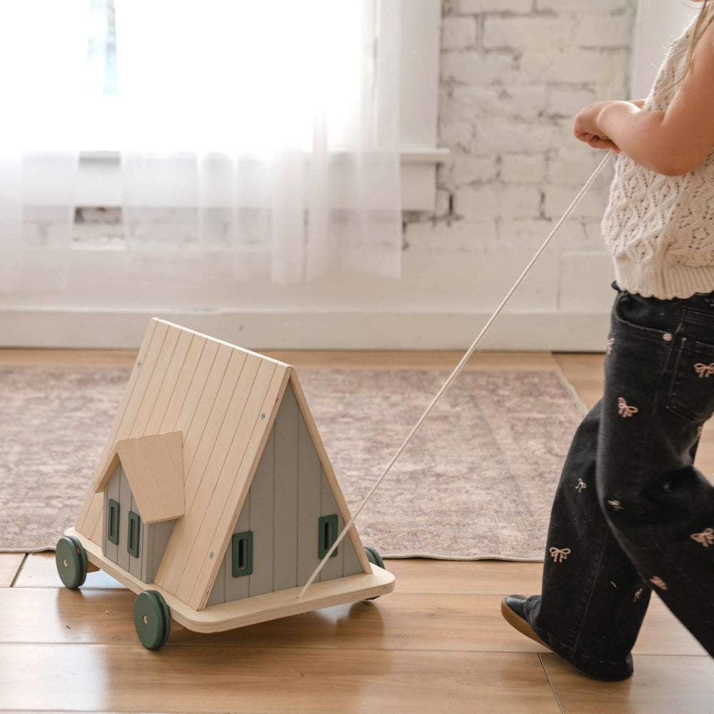 Person pulling a small wooden house toy on wheels across a wooden floor.