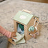 Toddler playing with wooden activity cube featuring green roof and colorful hanging toys on carpet
