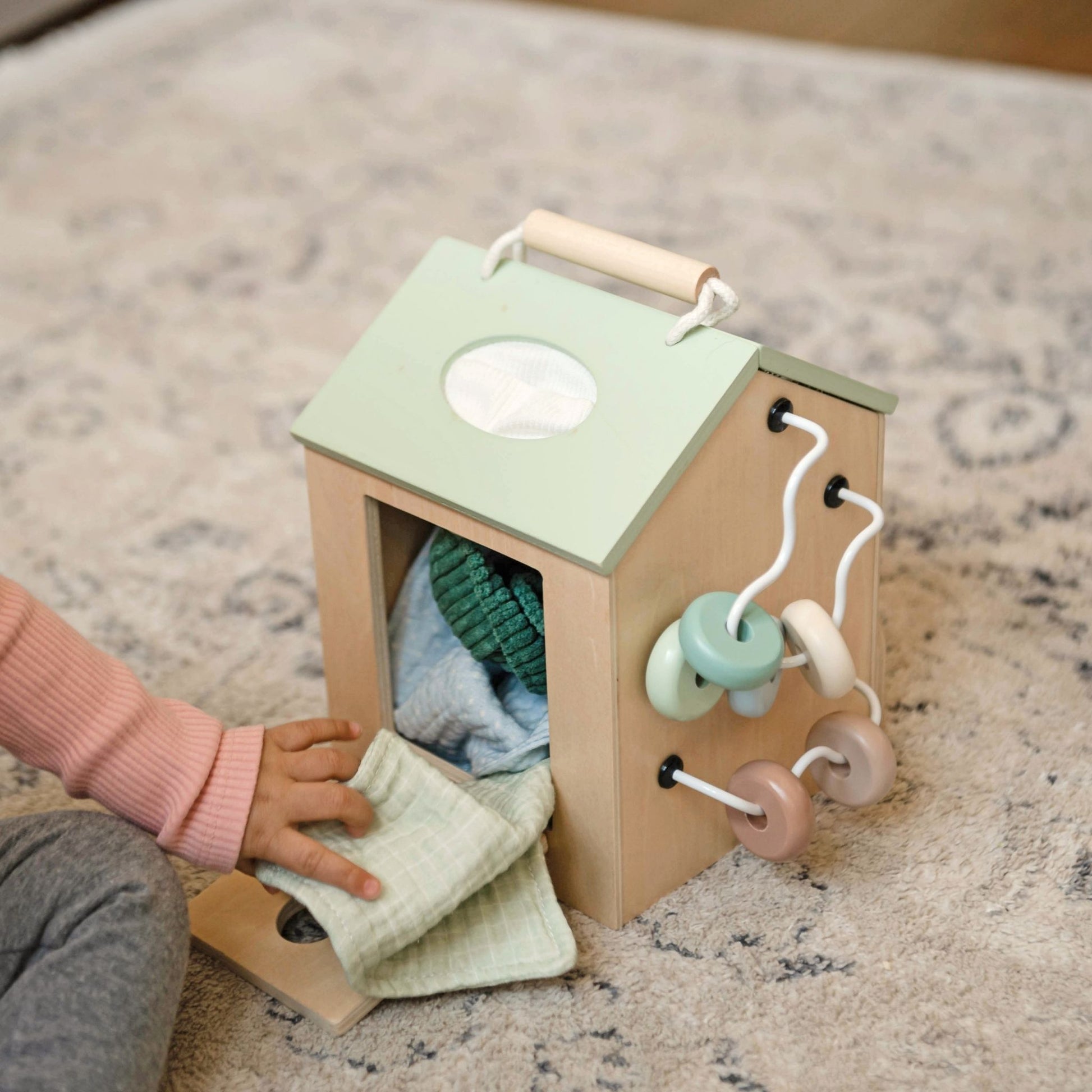 Toddler playing with wooden activity cube featuring green roof and colorful hanging toys on carpet