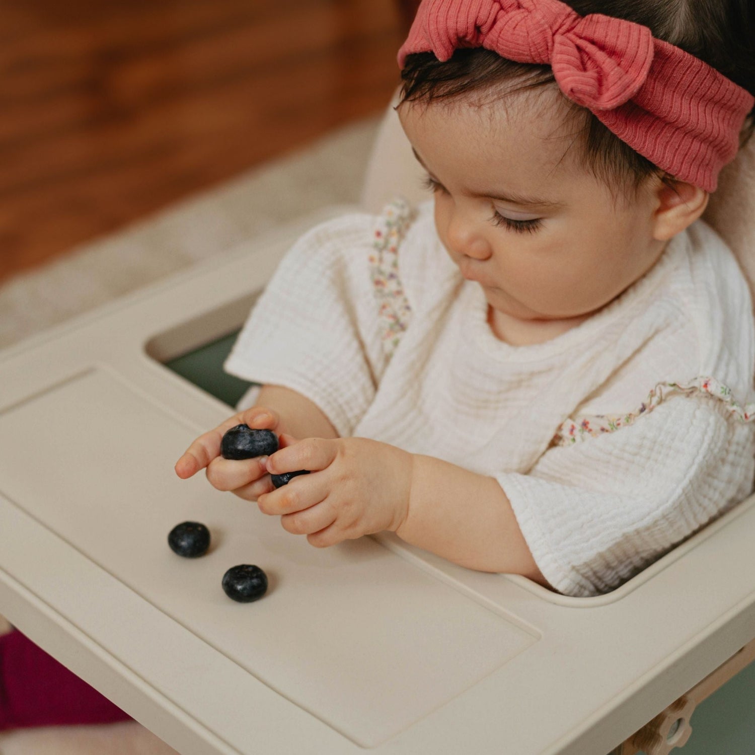 Baby sitting in a little wood chair holding blackberries and eating them from a dishwasher-safe silicone mat.