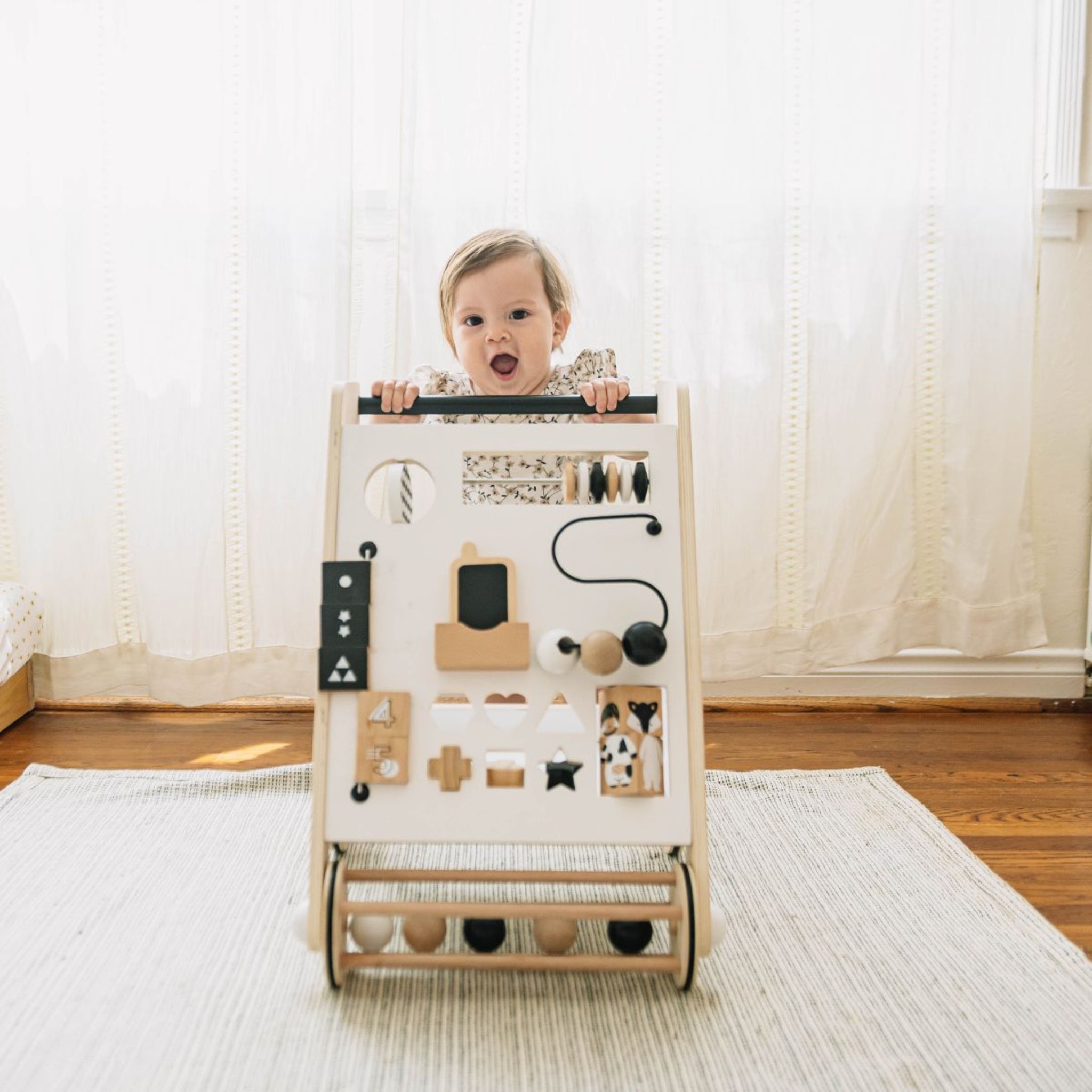Baby standing behind the Awesome Activity Walker on a woven rug by a sunlit window
