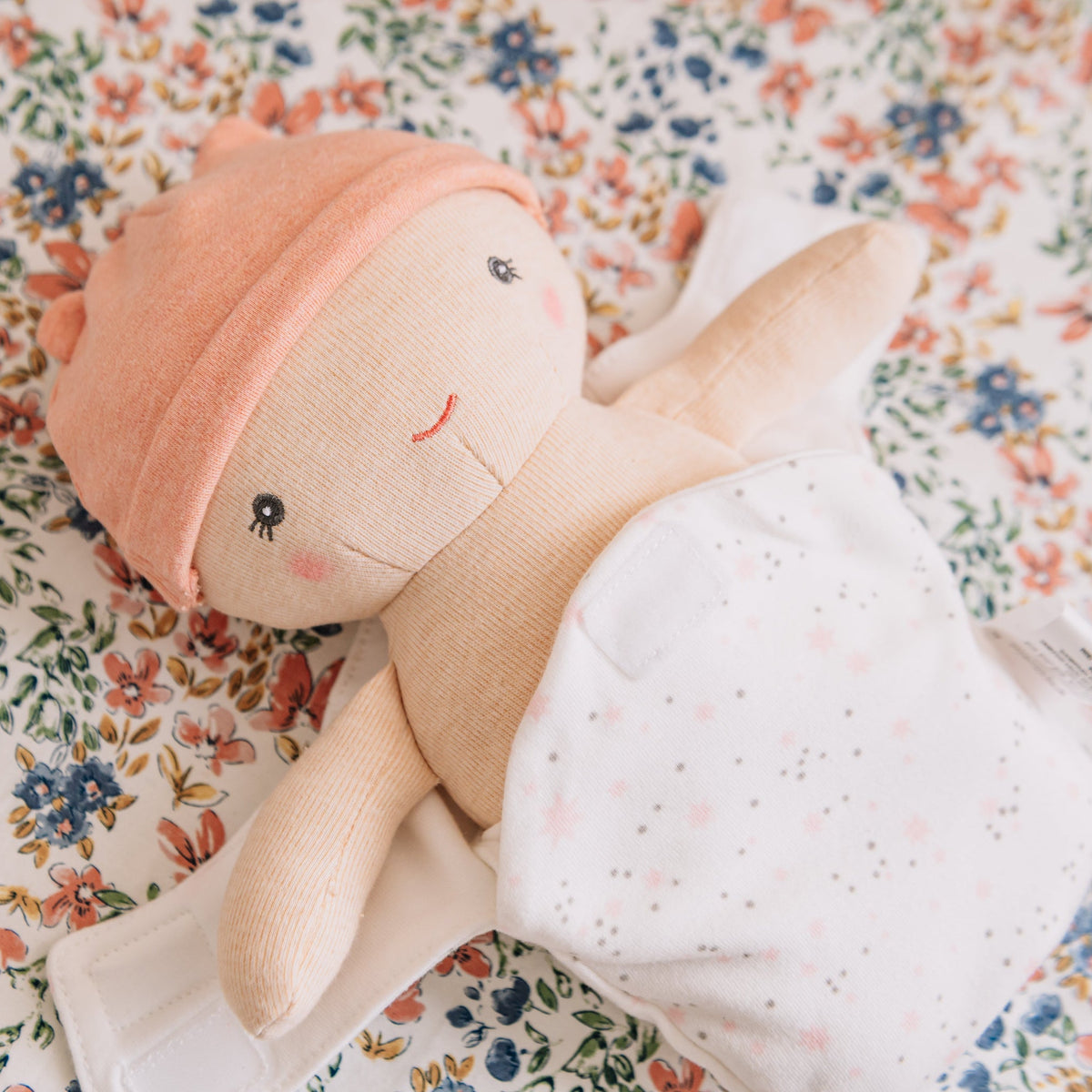 Close-up of the Cookie baby bundle doll's embroidered face and peach cap on a floral quilt.