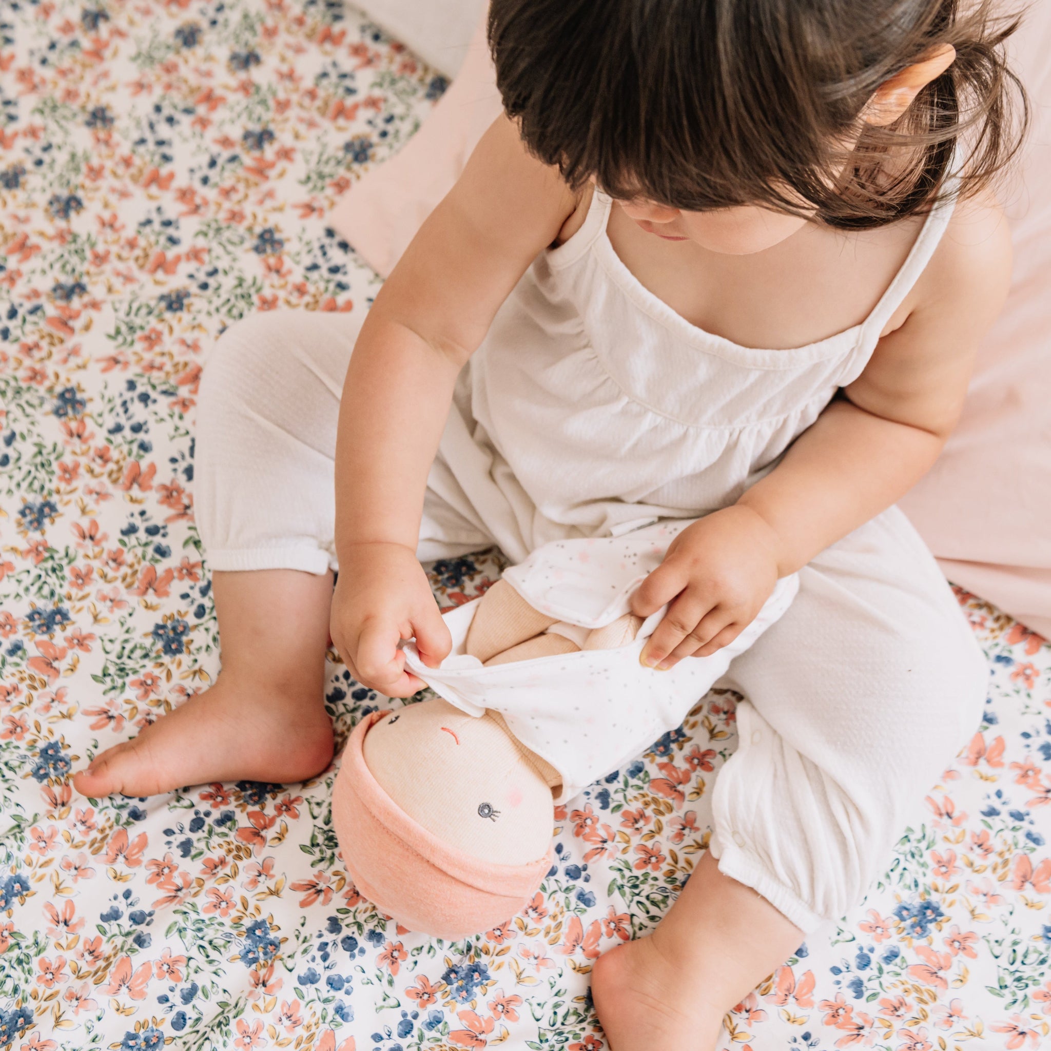 Toddler on a floral quilt swaddling the Cookie Baby Bundle Doll.
