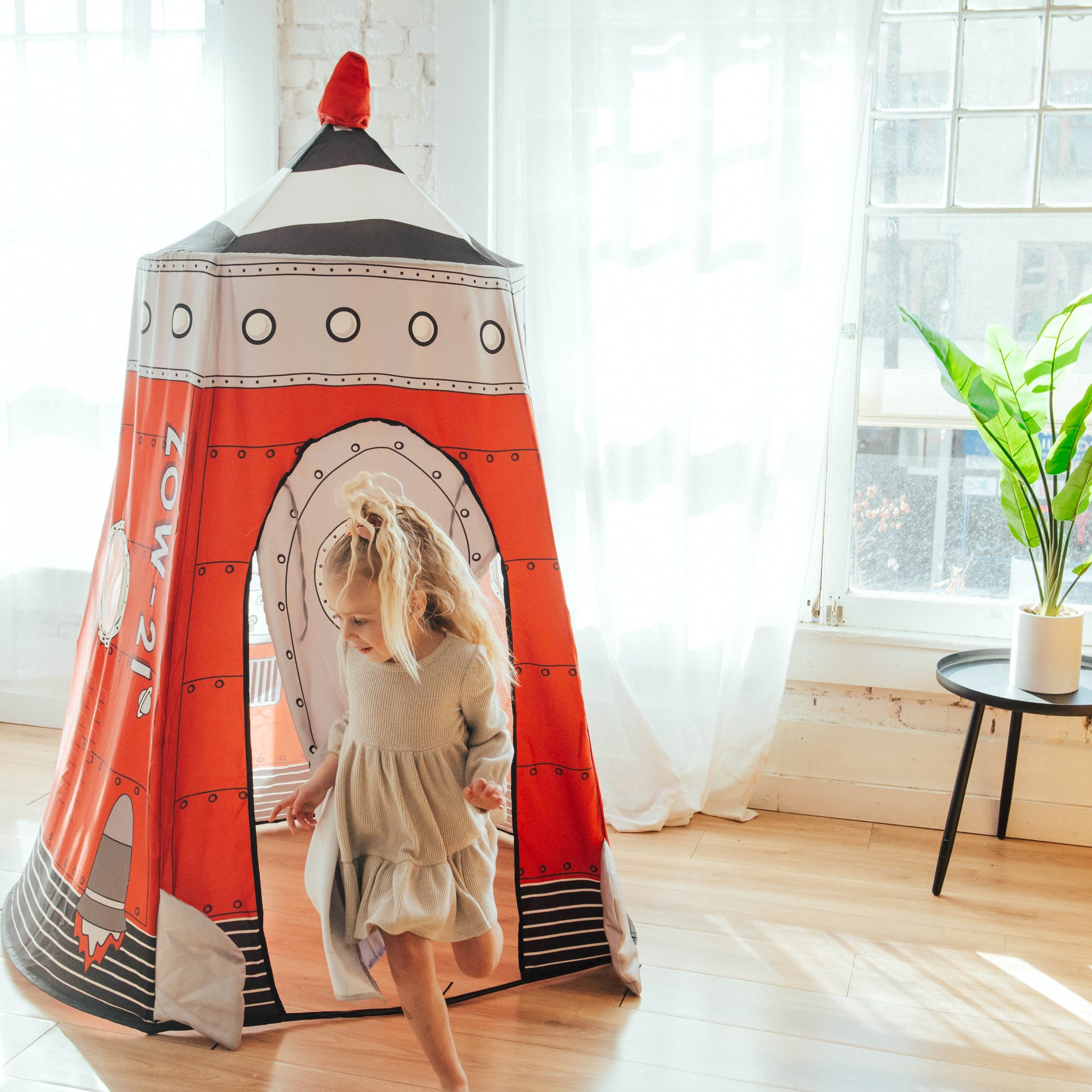 Child runs out through the front door of the red Rocket Pop-up Playhome, printed with porthole windows and a ZOW-21 rocket graphic