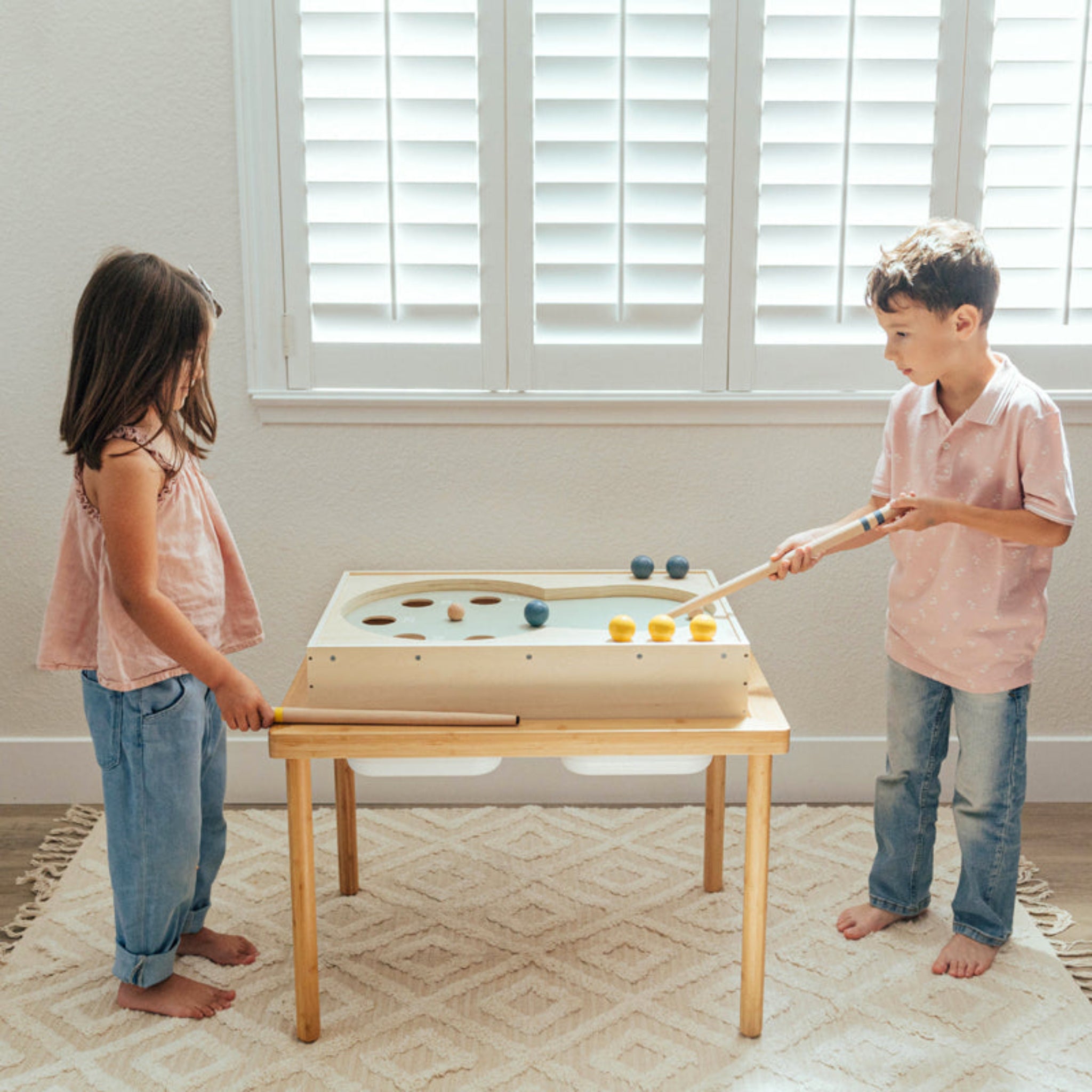 Two children playing with a small pool table in a room with a window.
