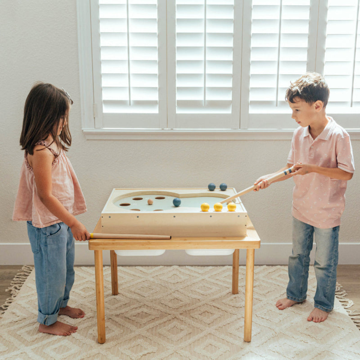 Two children playing with a small pool table in a room with a window.