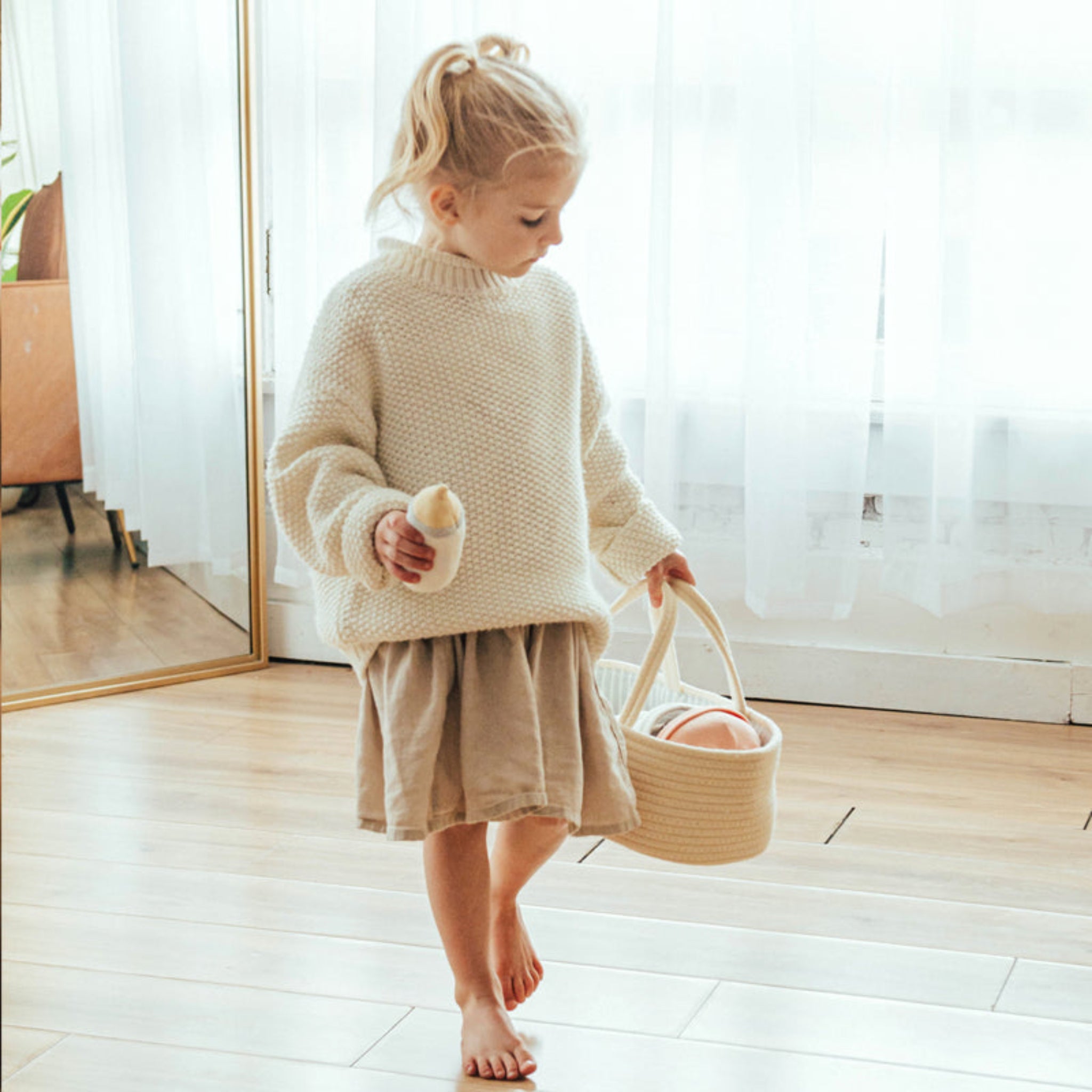 Child walks across a wooden floor carrying the Doll Carrier by its rope handles with a plush bottle in hand
