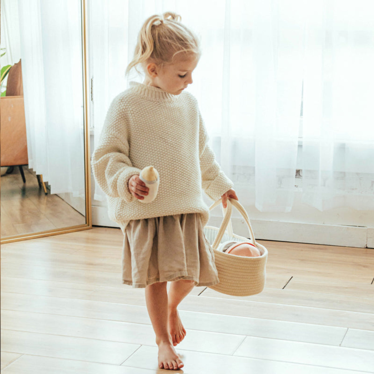 Child walks across a wooden floor carrying the Doll Carrier by its rope handles with a plush bottle in hand