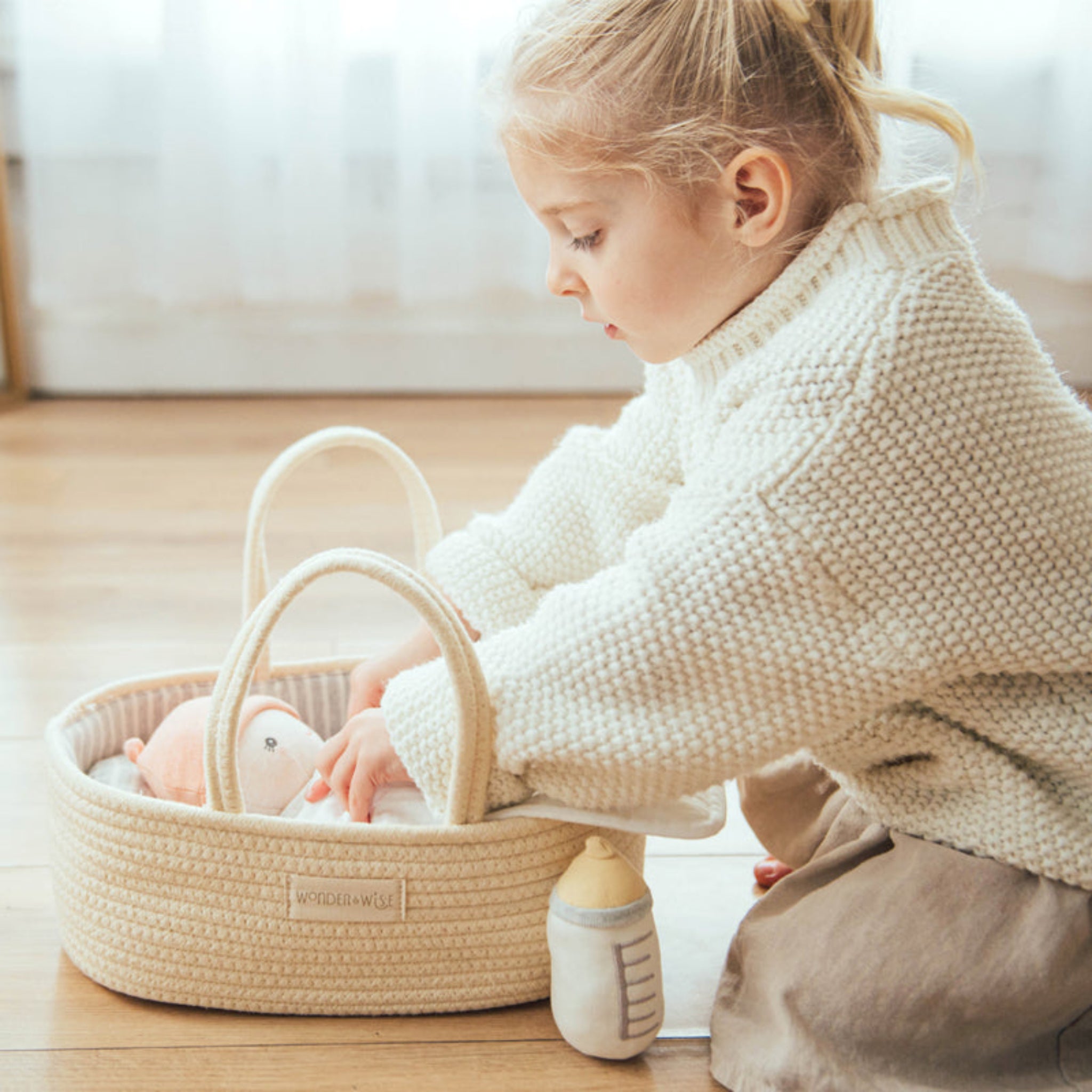 Child in a cream sweater tucks a doll into the Doll Carrier on a wooden floor, plush bottle beside it

