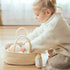 Child in a cream sweater tucks a doll into the Doll Carrier on a wooden floor, plush bottle beside it
