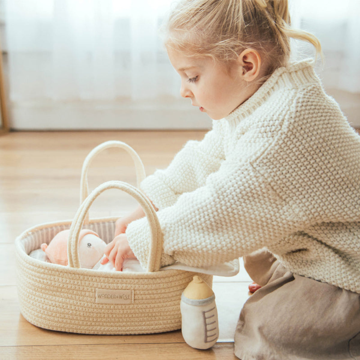Child in a cream sweater tucks a doll into the Doll Carrier on a wooden floor, plush bottle beside it