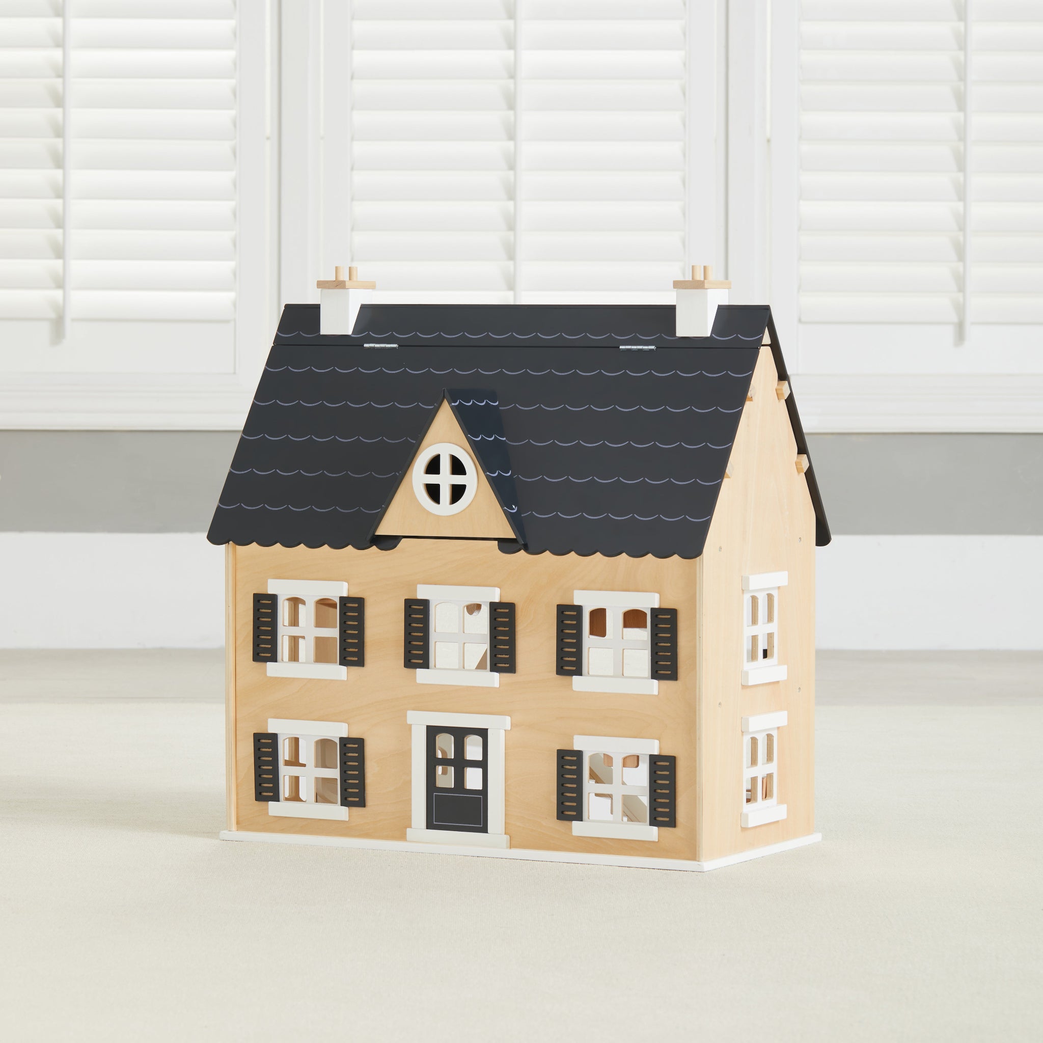 Wooden Dollhouse with black roof and white-trimmed shuttered windows, angled in front of white plantation shutters
