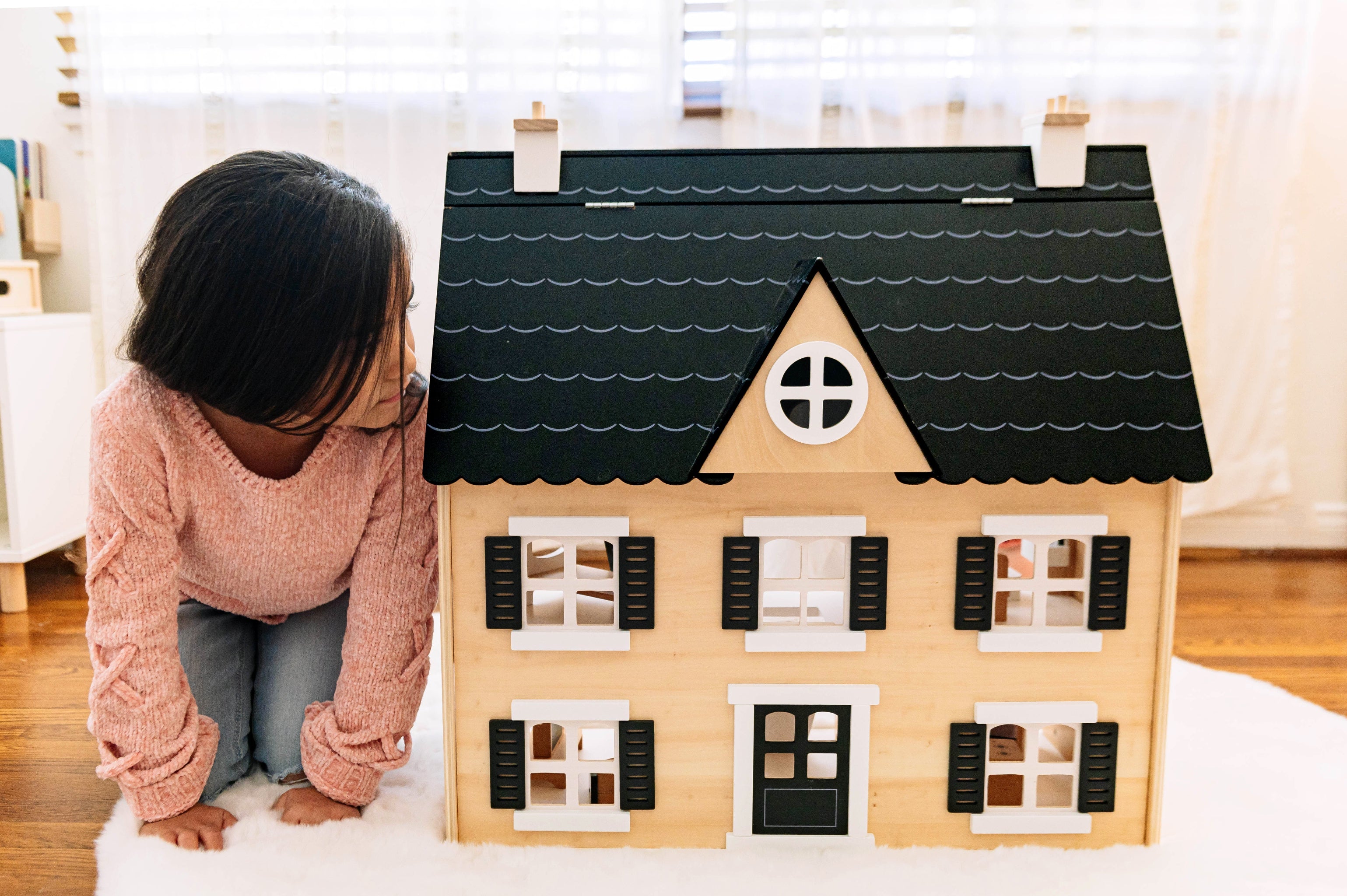 Child kneeling beside the wooden Dollhouse, looking at its black scalloped roof, dormer window and shuttered windows
