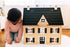 Child kneeling beside the wooden Dollhouse, looking at its black scalloped roof, dormer window and shuttered windows
