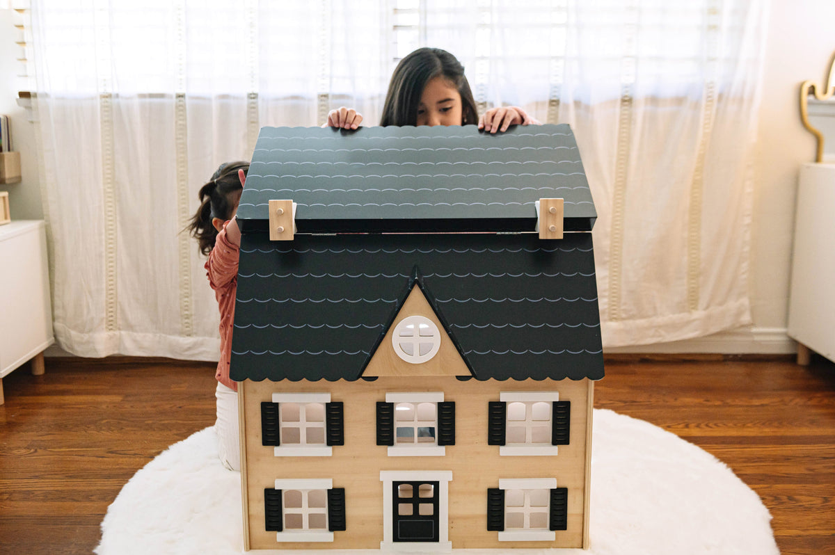 Two children lifting the hinged black roof of the wooden Dollhouse on a white rug in a sunlit bedroom