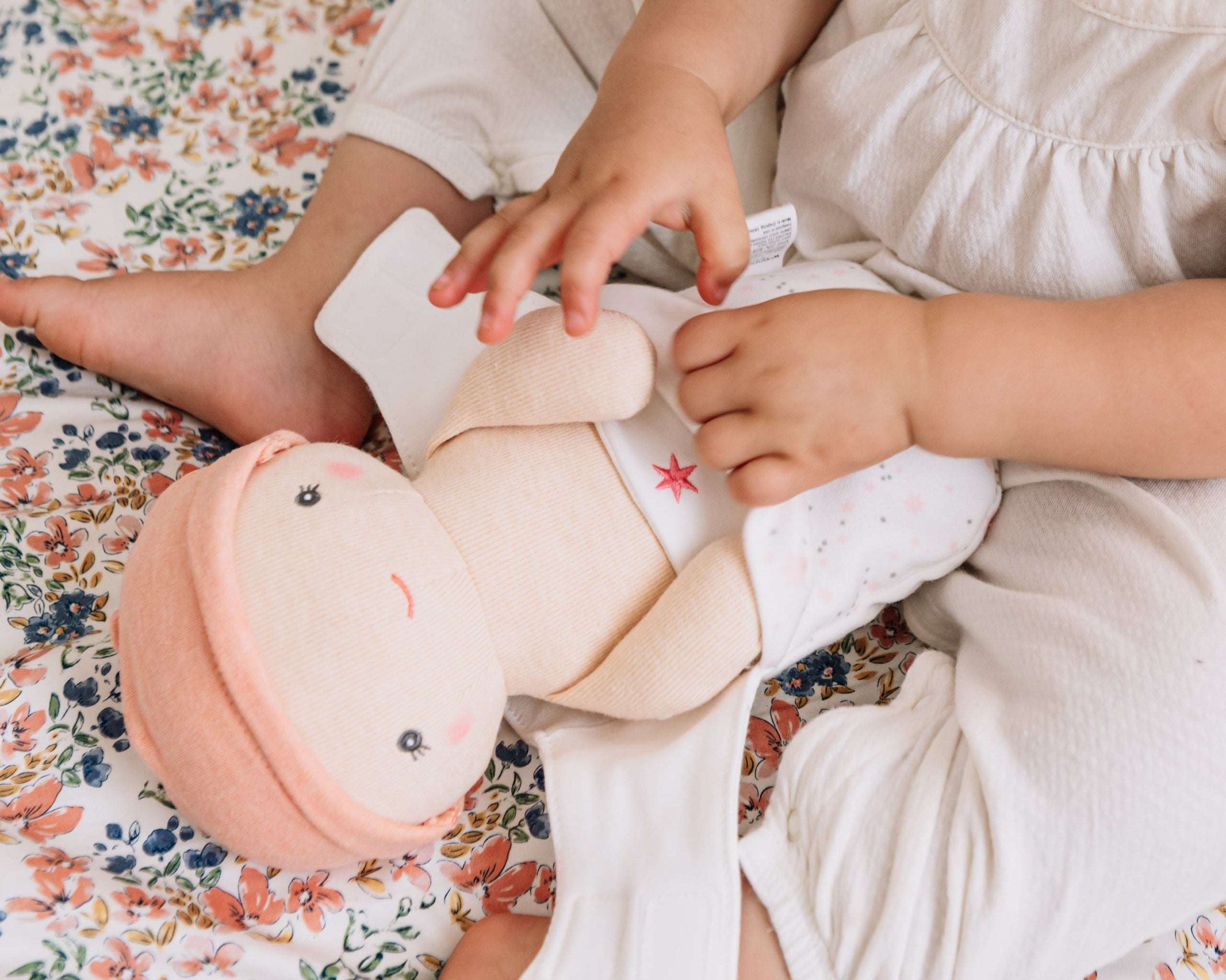 Toddler on a floral quilt unwrapping the Cookie doll that comes with the Dots and Rainbows Doll Carrier
