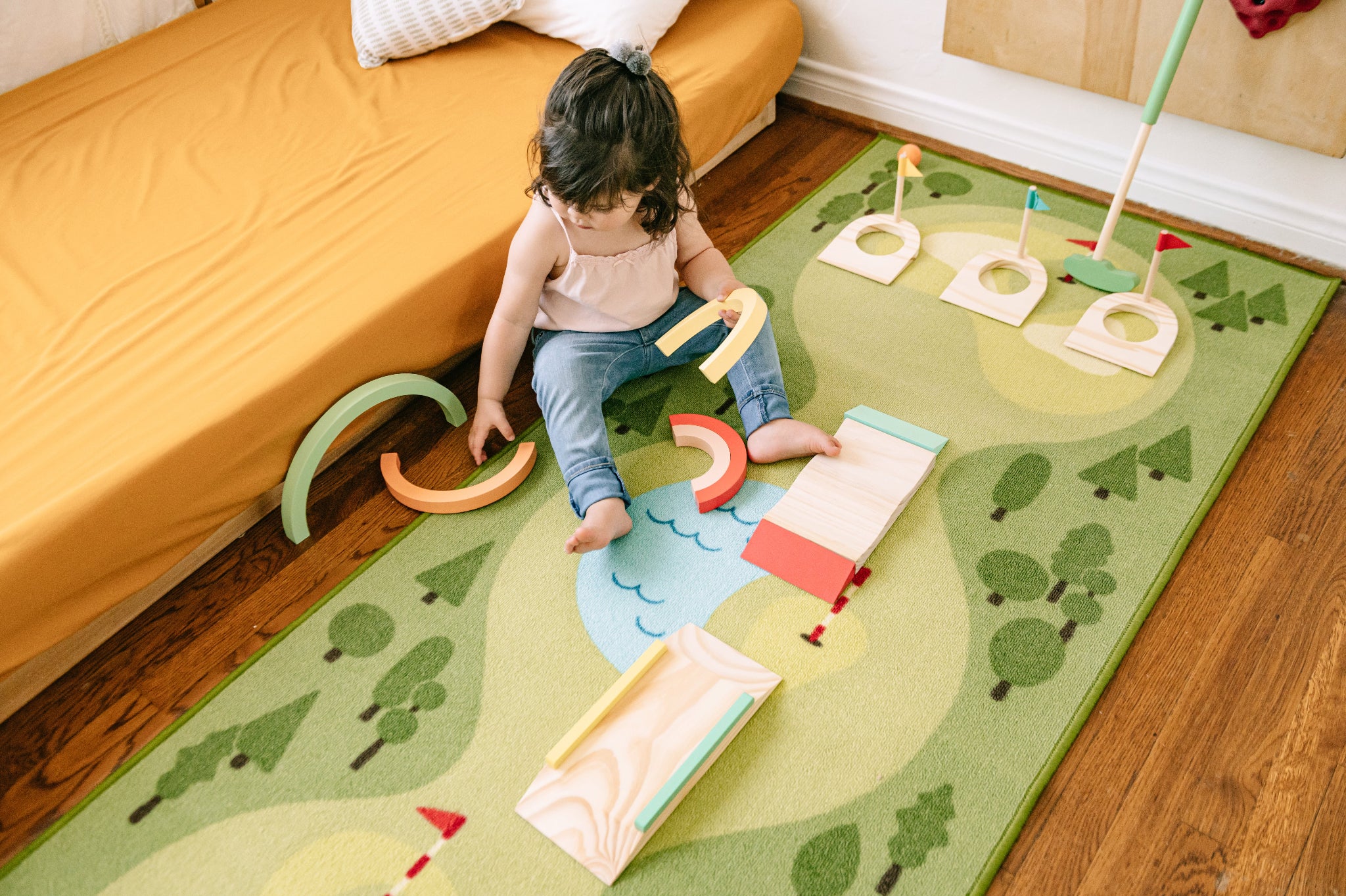 A toddler sits on a colorful children's rug in a bedroom, playing with wooden toy golf clubs and arches. The rug features a golf course design with trees, fairways, and a river, and is placed on a wooden floor next to a bed with an orange sheet.