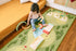 A toddler sits on a colorful children's rug in a bedroom, playing with wooden toy golf clubs and arches. The rug features a golf course design with trees, fairways, and a river, and is placed on a wooden floor next to a bed with an orange sheet.