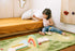 A young child sits on a green patterned rug in a child's bedroom, playing with wooden books and a rainbow toy near a bed with mustard yellow bedding.