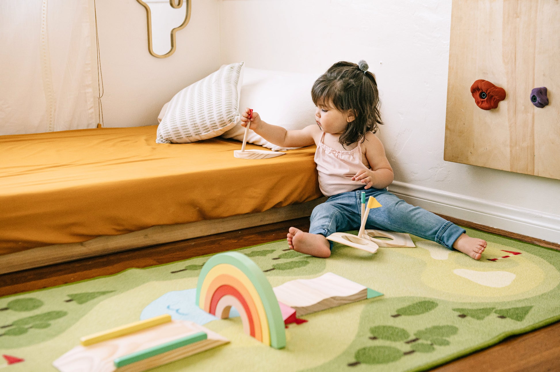 A young child sits on a green patterned rug in a child's bedroom, playing with wooden books and a rainbow toy near a bed with mustard yellow bedding.