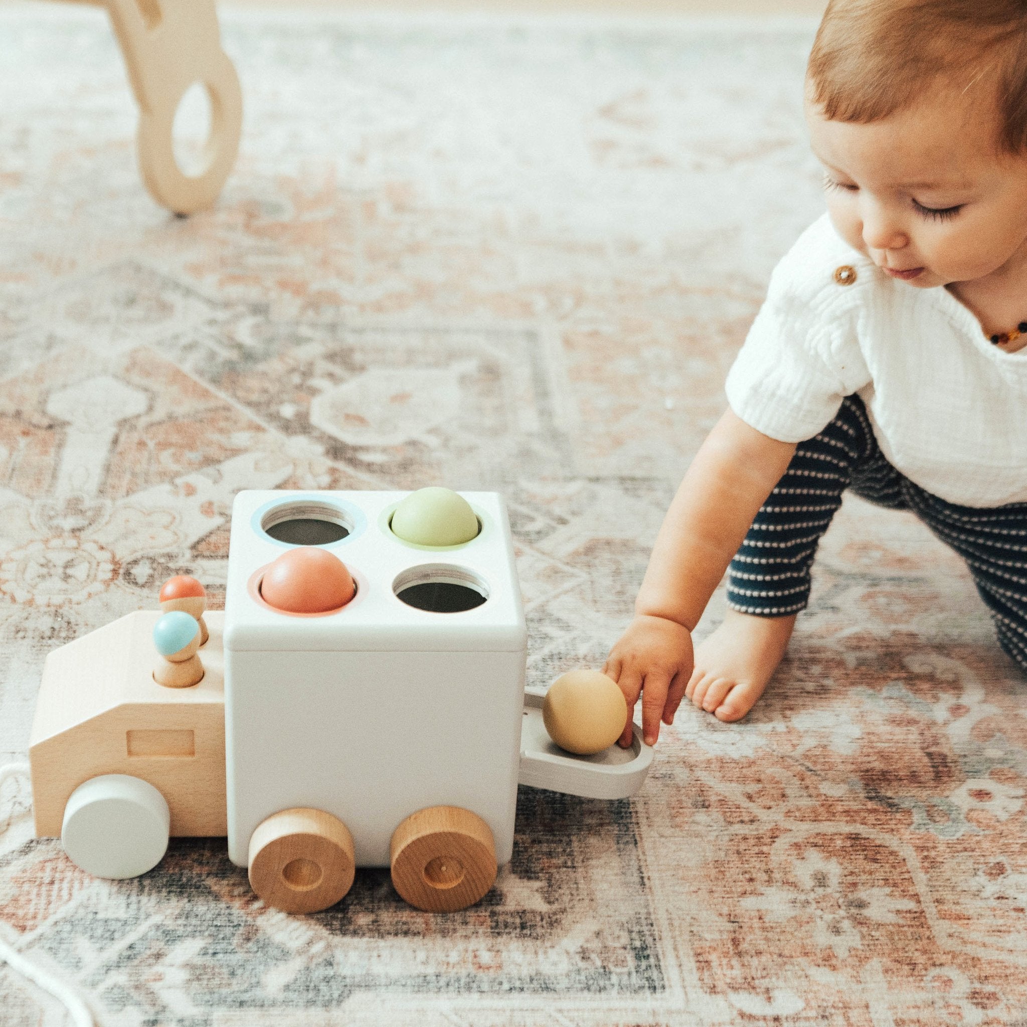 Baby on a patterned rug pulling a wooden ball out of the rear door of the Hammer Time Pull Toy
