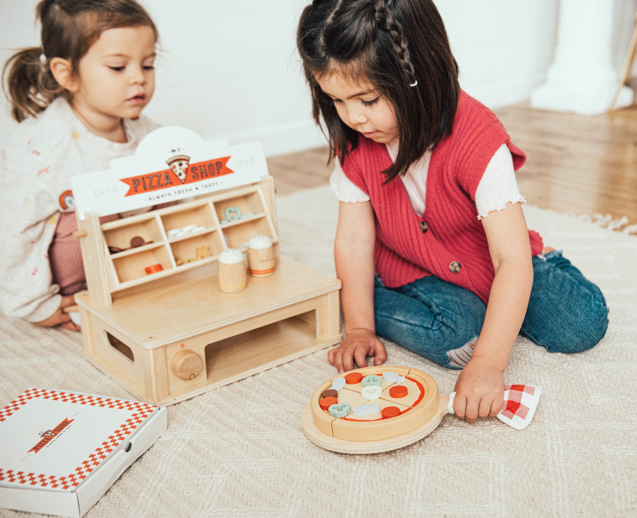 Two girls kneel on a cream rug playing with the Wonder & Wise My Pizza Oven: a wooden pizza shop counter with a “My Pizza Shop” sign and topping cubbies, a wooden pizza on a peel with sliced toppings, and a checkered pizza box.
