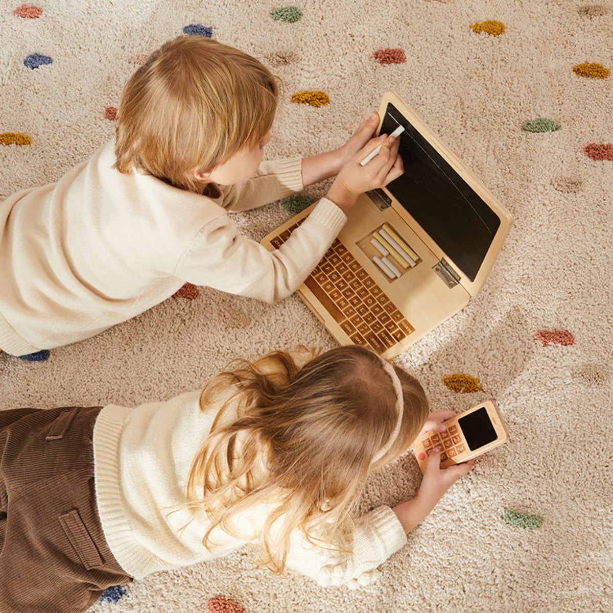 Overhead view of two children on a rug, one writing with chalk on the Nonstop Laptop screen, one holding the phone