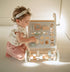A baby in a cream ruffle dress and pink pants plays with a wooden activity walker. The walker has a light gray front panel with various wooden shape-sorting shapes, gears, and a small car, along with a wooden roller bar with pastel-colored blocks at the bottom, set in a bright indoor environment.