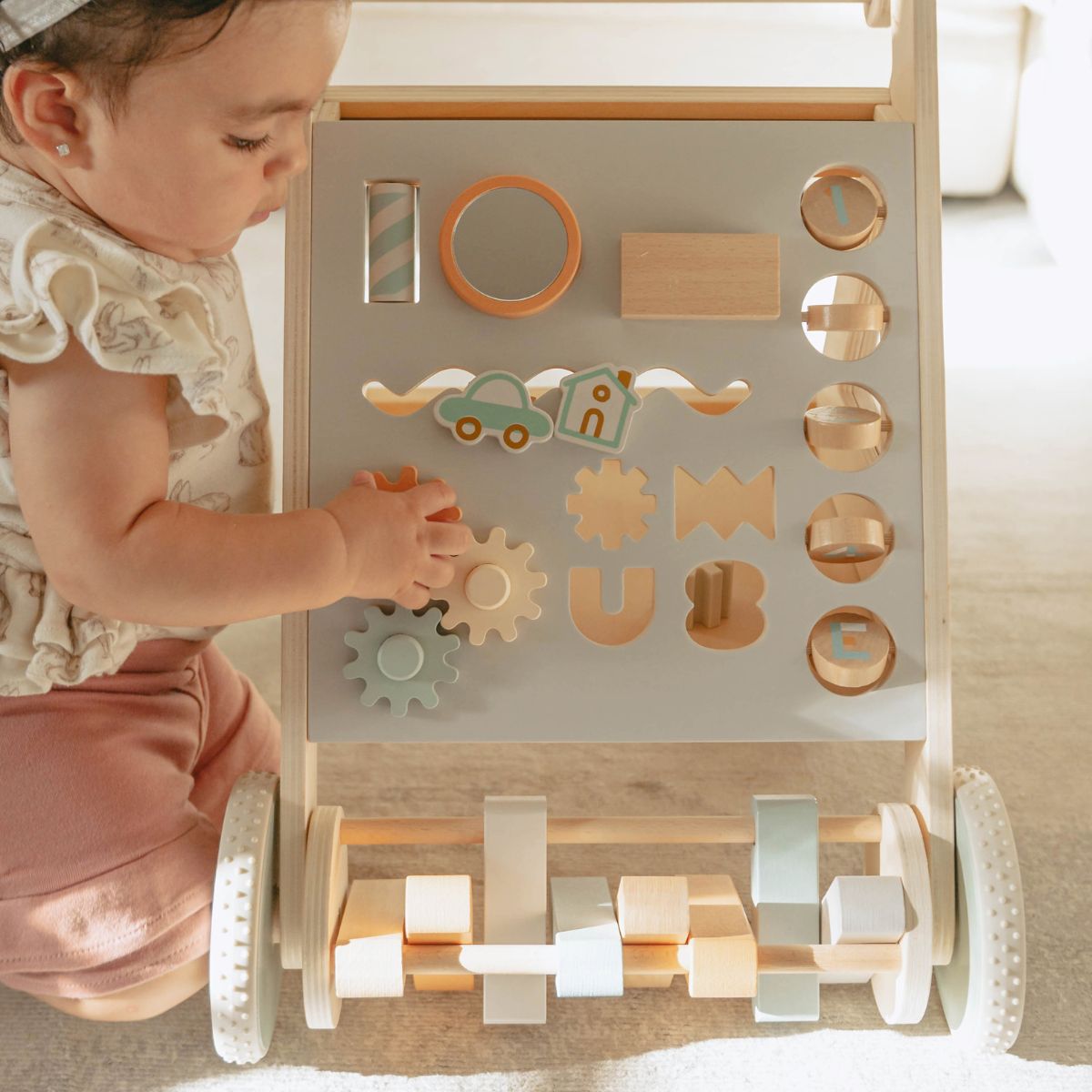 A baby in a cream ruffle dress and pink pants plays with a wooden activity walker. The walker has a light gray front panel with various wooden shape-sorting shapes, gears, and a small car, along with a wooden roller bar with pastel-colored blocks at the bottom, set in a bright indoor environment.