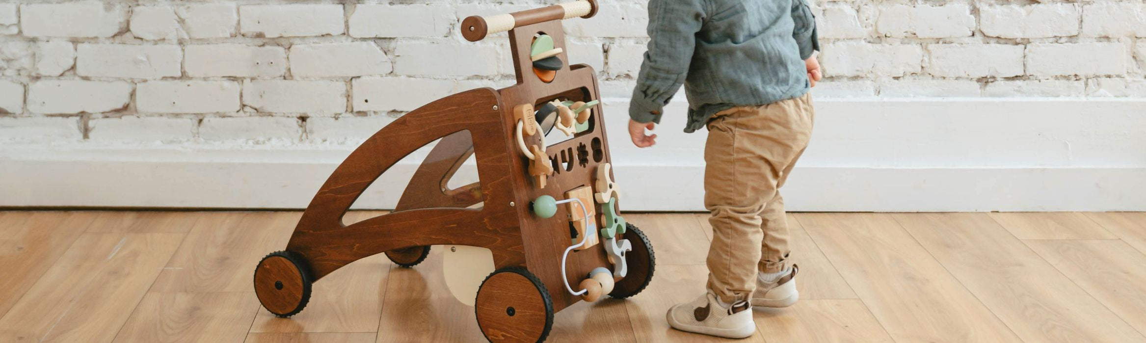 A toddler wearing a blue jacket and tan pants walks beside a wooden activity walker toy with wheels and hanging toys in a bright room with a white brick wall and wood floor.