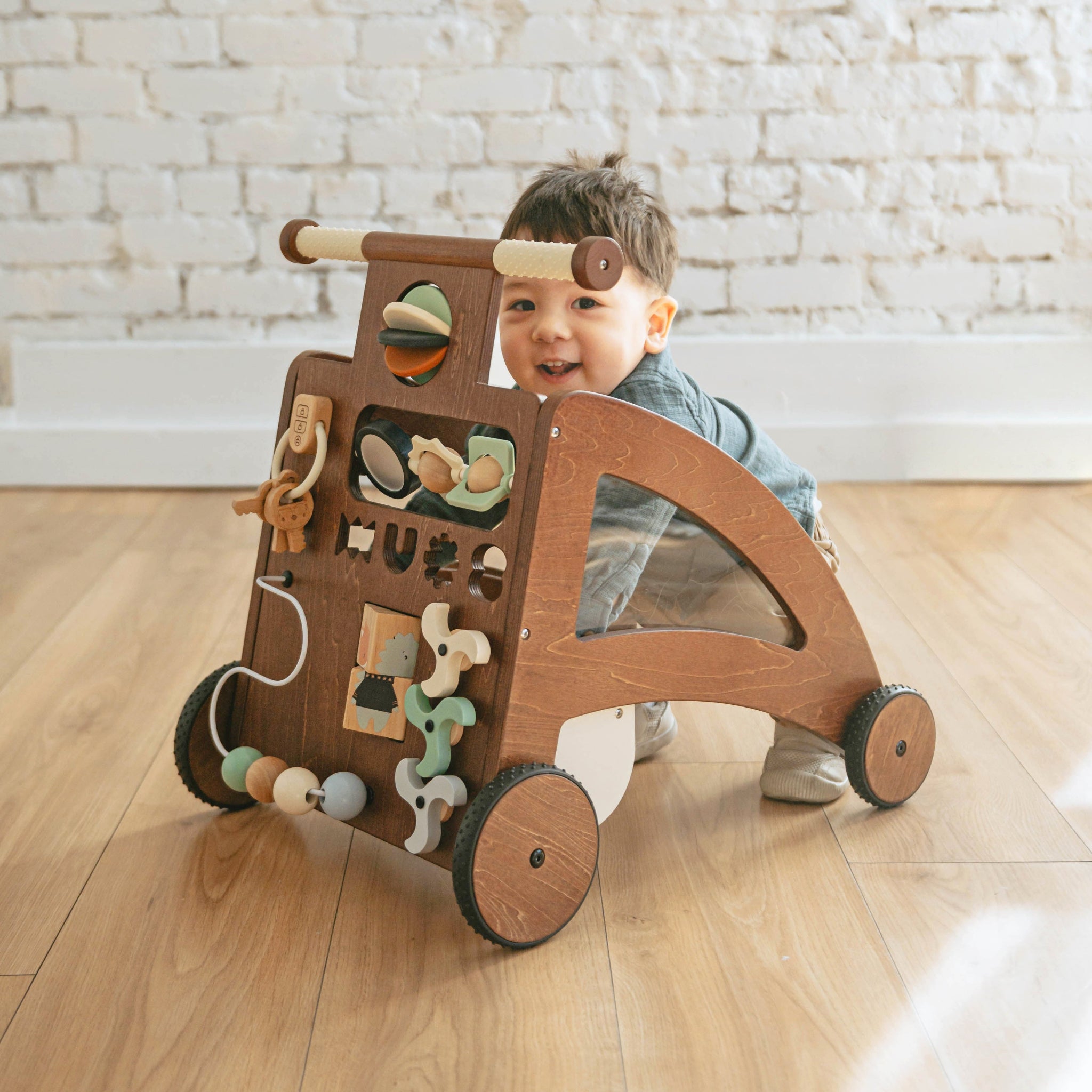 Toddler smiles from behind the Waldo Activity Walker, a dark wood push walker with an activity front, on a wood floor
