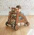 Toddler smiles from behind the Waldo Activity Walker, a dark wood push walker with an activity front, on a wood floor