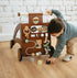 Toddler holds the handle of the Waldo Activity Walker and reaches for blocks on the floor by a white brick wall