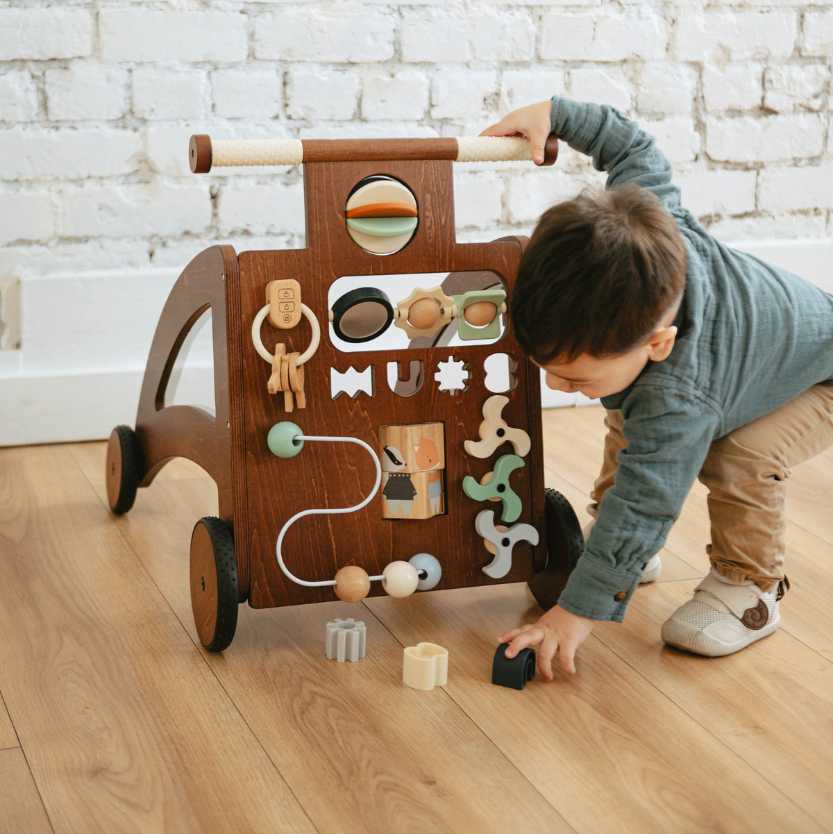 Toddler holds the handle of the Waldo Activity Walker and reaches for blocks on the floor by a white brick wall