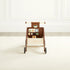 Back  view of the Waldo Activity Walker, a walnut-stained wooden push walker with a cream handle, an activity panel with a mirror, spinners, and shape toys, a storage bin, and black rubber wheels, in a white paneled room.