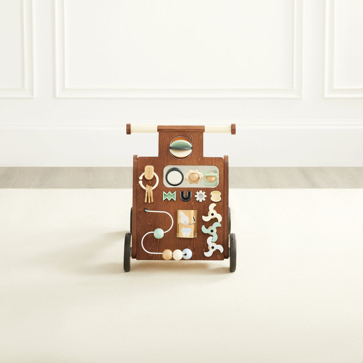 Front  view of the Waldo Activity Walker, a walnut-stained wooden push walker with a cream handle, an activity panel with a mirror, spinners, and shape toys, a storage bin, and black rubber wheels, in a white paneled room.