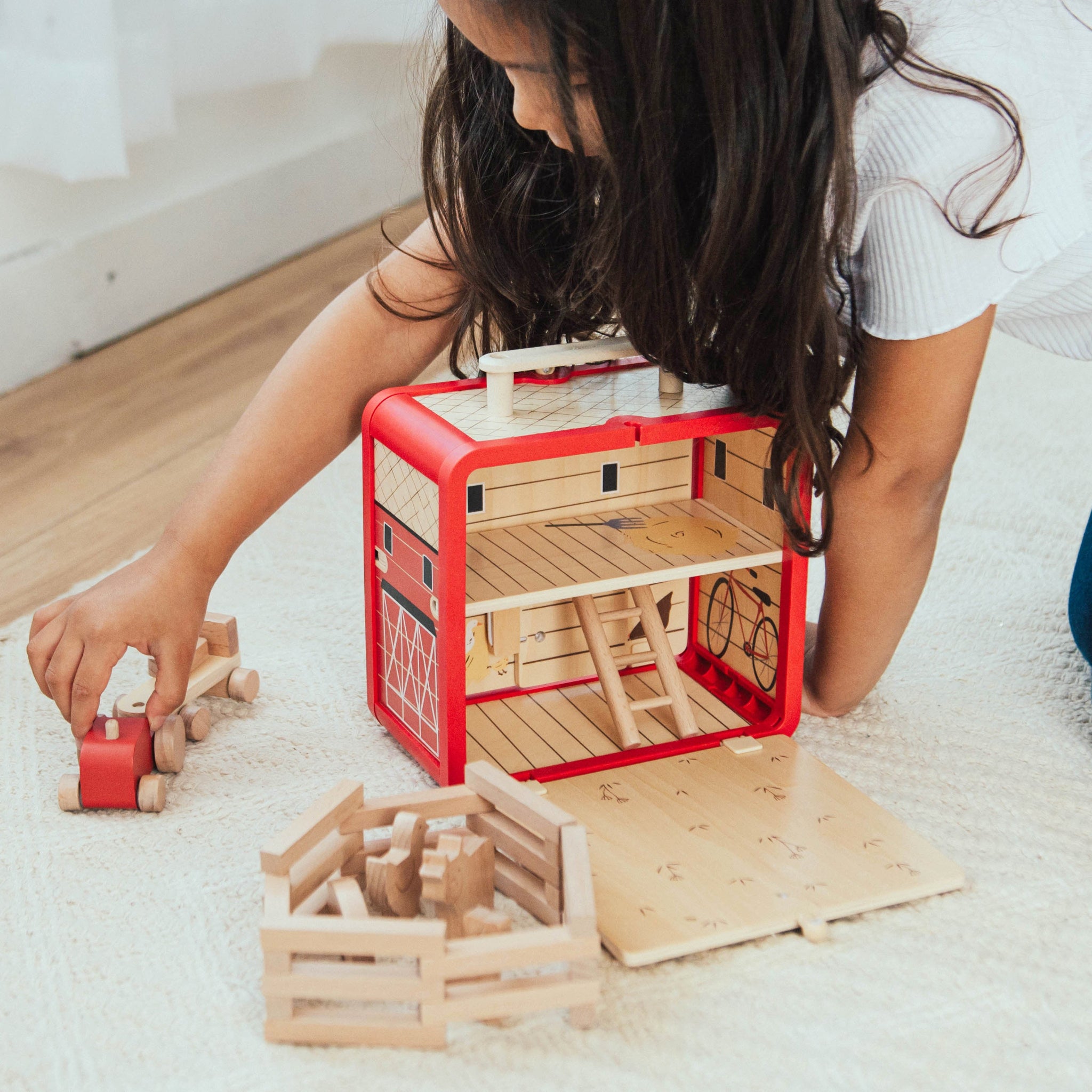 A girl kneels on a cream rug playing with the open Take with Me Farm, a red wooden barn with a hayloft and ladder inside, a wooden fence pen, and a red tractor she pushes beside it.