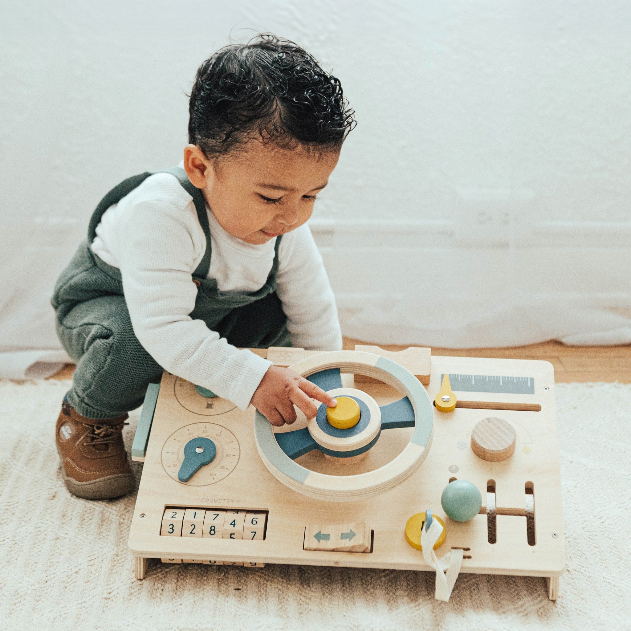 Baby kneeling on a rug pressing the yellow horn at the center of the Beep! Beep! Busy Board steering wheel