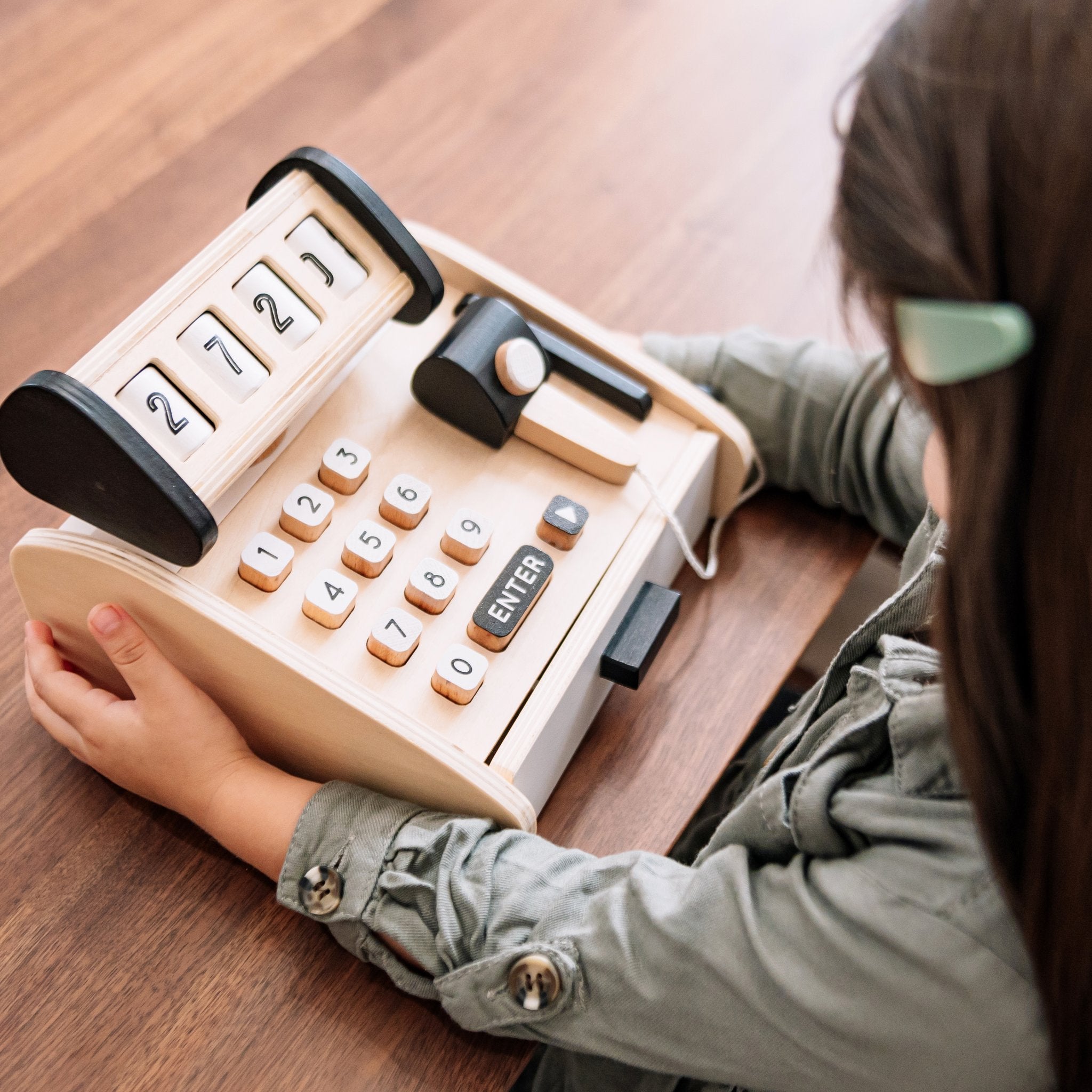 Child pressing the number keys on the wooden Cash Register at a wood table, display reading 2721