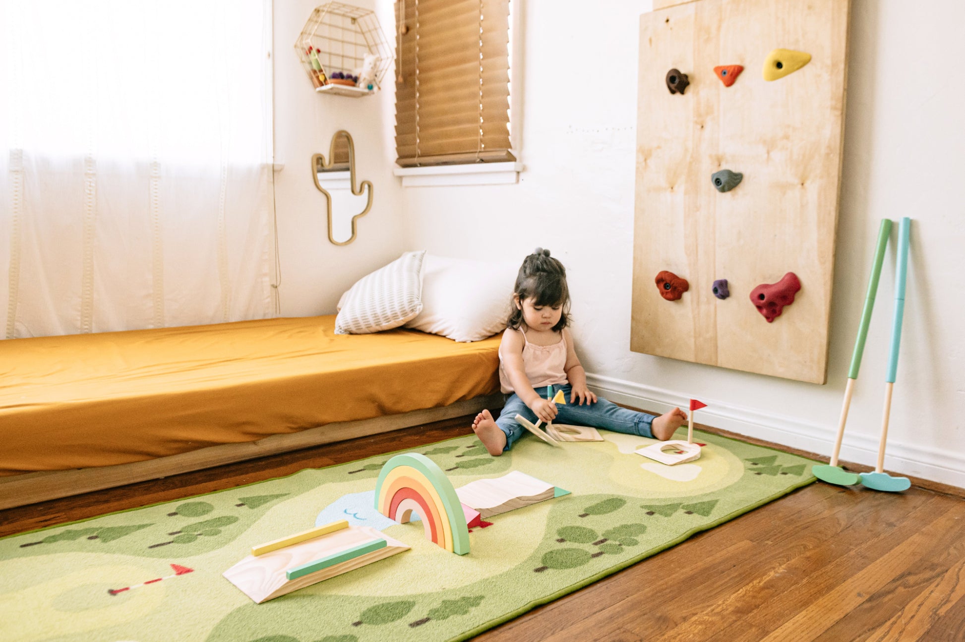 A young child sits on a green green golf mat modern playroom, surrounded by toys including a rainbow toy, a small golf set, and a wooden climbing wall.
